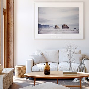 May include: A white couch with a throw blanket and a vase of white branches on a coffee table with two books and a brown ceramic vase. A framed photograph of a beach scene with two rock formations hangs on the wall above the couch.