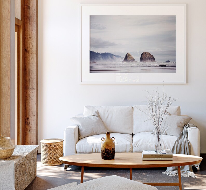 May include: A white couch with a throw blanket and a vase of white branches on a coffee table with two books and a brown ceramic vase. A framed photograph of a beach scene with two rock formations hangs on the wall above the couch.
