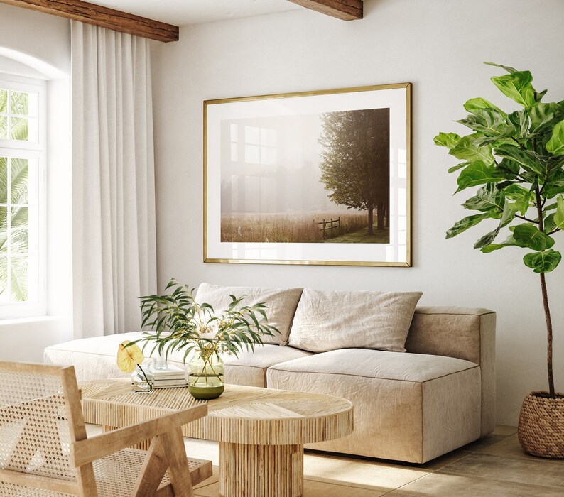 K&ouml;nnte beinhalten: Ein hellbeiges Ecksofa mit zwei Kissen steht in einem Wohnzimmer mit einem gro&szlig;en gerahmten Druck einer nebligen Landschaft dar&uuml;ber. Ein kleiner Holz-Couchtisch steht vor dem Sofa, und eine Topfpflanze mit gro&szlig;en gr&uuml;nen Bl&auml;ttern steht in der Ecke des Raumes.