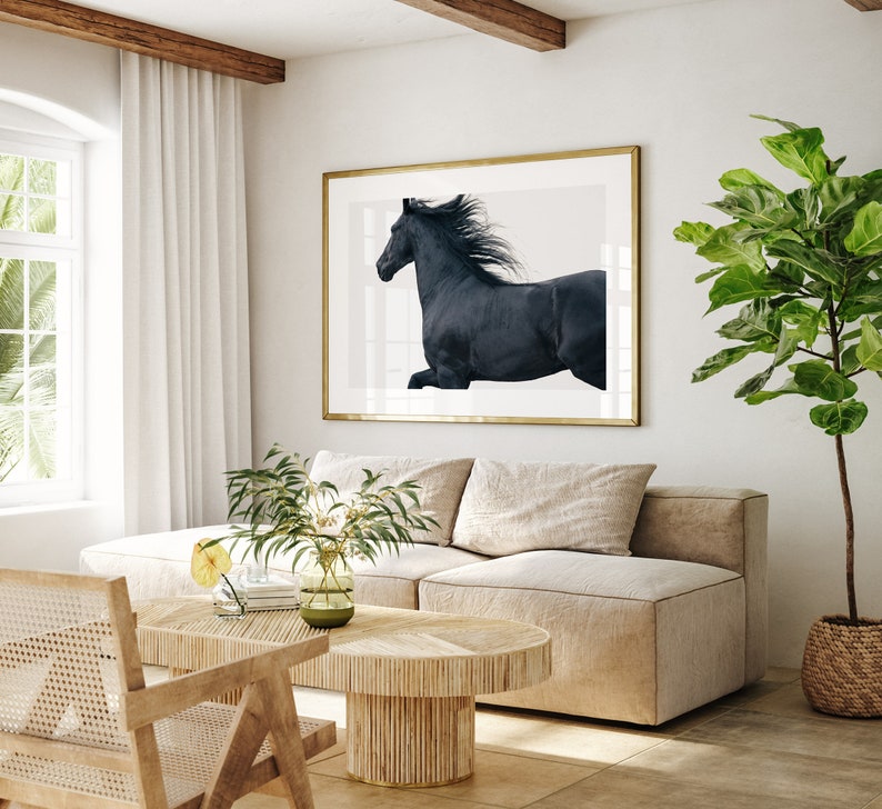 May include: A black horse with a flowing mane runs across a white background in a framed print. The print is hanging on a white wall in a living room setting with a beige sofa and a wooden coffee table.