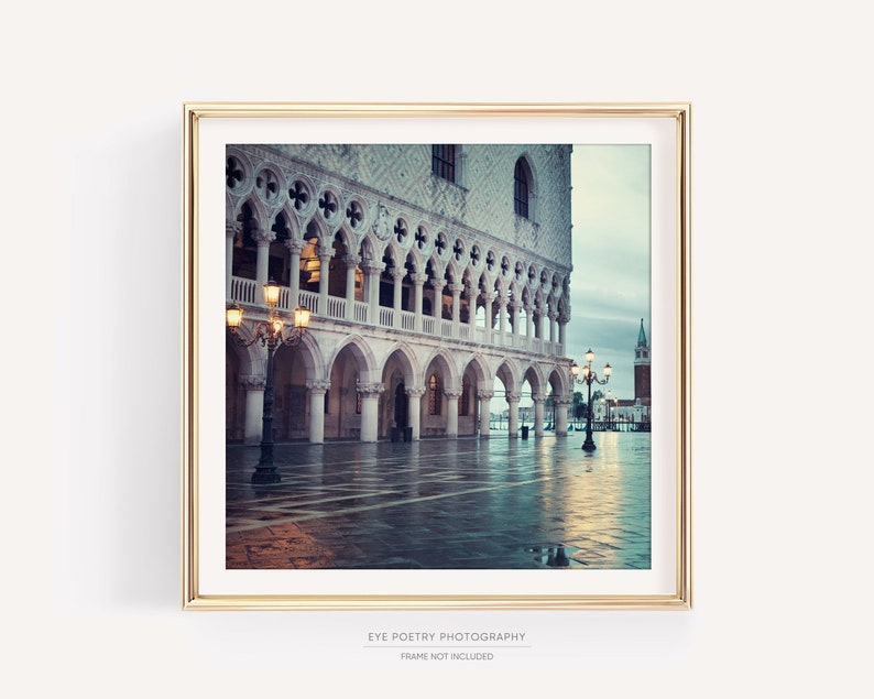 May include: A photograph of a building with arches and columns in Venice, Italy. The building is lit by streetlights and the ground is wet from rain. The photo is framed in a gold frame.