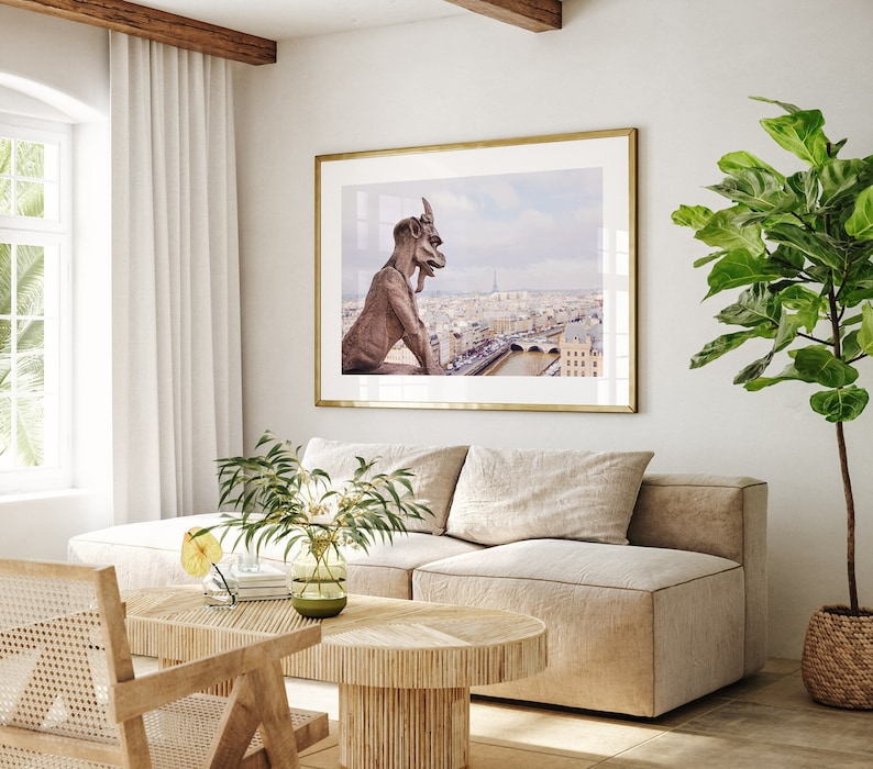 May include: A living room interior with a beige sofa, a wooden coffee table, and a framed print of a gargoyle overlooking a cityscape. A potted plant with green leaves is in the corner of the room.