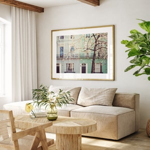 May include: A framed photograph of a row of pastel green houses with a blooming tree in front. The houses have white trim and windows. The photo is hanging on a white wall above a beige couch.