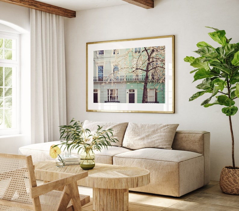 May include: A framed photograph of a row of pastel green houses with a blooming tree in front. The houses have white trim and windows. The photo is hanging on a white wall above a beige couch.