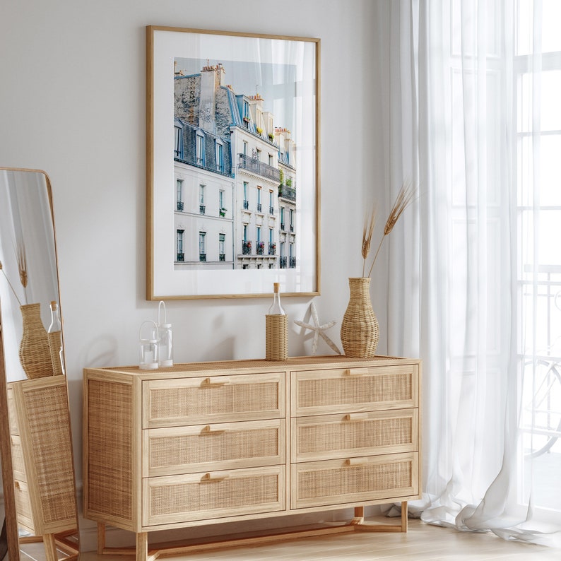 Puede incluir: Una c&oacute;moda de seis cajones con un frente de rat&aacute;n tejido y patas de madera. La c&oacute;moda est&aacute; decorada con una impresi&oacute;n enmarcada de un edificio parisino, un jarr&oacute;n con hierbas secas y una botella de vidrio con una estrella de mar.