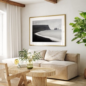 May include: A living room interior with a beige sectional sofa, a wooden coffee table, a potted plant, and a framed black and white photograph of a coastal landscape.