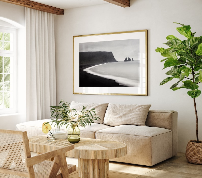 May include: A living room interior with a beige sectional sofa, a wooden coffee table, a potted plant, and a framed black and white photograph of a coastal landscape.