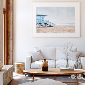 Peut inclure: Un canap&eacute; blanc avec un plaid et un vase de branches blanches se trouve devant une photographie encadr&eacute;e d'une cabine de sauveteur bleue sur une plage.