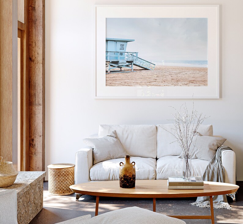 Peut inclure: Un canap&eacute; blanc avec un plaid et un vase de branches blanches se trouve devant une photographie encadr&eacute;e d'une cabine de sauveteur bleue sur une plage.