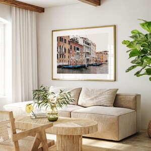 May include: A living room interior with a beige sectional sofa, a wooden coffee table, and a framed photograph of a canal in Venice, Italy. The photograph shows colorful buildings lining the canal, with gondolas floating in the water.