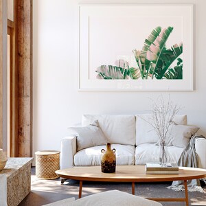 K&ouml;nnte beinhalten: Ein wei&szlig;es Sofa mit zwei Kissen und einer Wolldecke steht in einem Wohnzimmer mit einem Holz-Couchtisch. Eine Vase mit wei&szlig;en Zweigen und eine braune Keramikvase stehen auf dem Tisch. Ein gerahmtes Bild mit gr&uuml;nen und rosafarbenen Bananenbl&auml;ttern h&auml;ngt an der Wand.