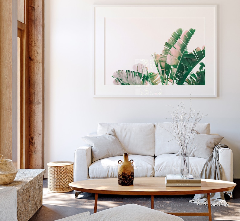 K&ouml;nnte beinhalten: Ein wei&szlig;es Sofa mit zwei Kissen und einer Wolldecke steht in einem Wohnzimmer mit einem Holz-Couchtisch. Eine Vase mit wei&szlig;en Zweigen und eine braune Keramikvase stehen auf dem Tisch. Ein gerahmtes Bild mit gr&uuml;nen und rosafarbenen Bananenbl&auml;ttern h&auml;ngt an der Wand.