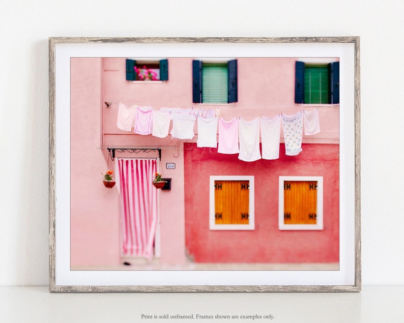 Peut inclure: Un b&acirc;timent rose avec un auvent ray&eacute; au-dessus de la porte et du linge suspendu &agrave; une corde &agrave; linge. Les fen&ecirc;tres sont vertes et les volets sont bleus. Il y a deux plantes en pot accroch&eacute;es au mur.