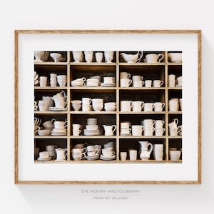 May include: A white wooden shelf unit filled with white ceramic mugs, cups, bowls, and pitchers. The shelf unit is against a white wall.