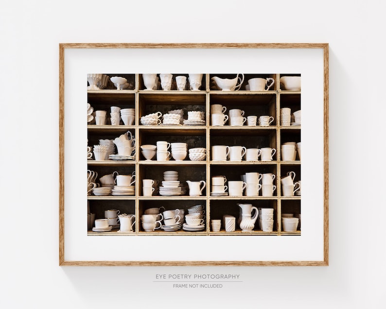 May include: A white wooden shelf unit filled with white ceramic mugs, cups, bowls, and pitchers. The shelf unit is against a white wall.