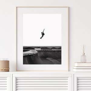 Peut inclure: Tirage encadr&eacute; en noir et blanc d'un skateboarder en plein saut au-dessus d'un bowl de skatepark. La photo capture la silhouette du skateur contre un ciel blanc, soulignant l'action dynamique et les courbes de la structure en b&eacute;ton.