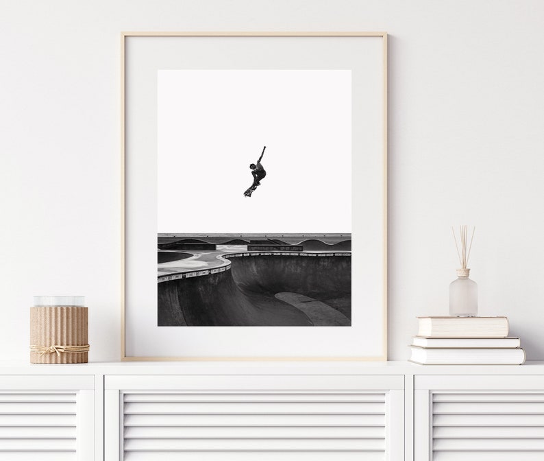 Peut inclure: Tirage encadr&eacute; en noir et blanc d'un skateboarder en plein saut au-dessus d'un bowl de skatepark. La photo capture la silhouette du skateur contre un ciel blanc, soulignant l'action dynamique et les courbes de la structure en b&eacute;ton.