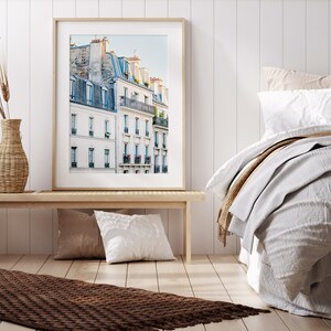 Puede incluir: Una fotograf&iacute;a enmarcada de un edificio parisino con paredes blancas y ventanas azules. El edificio tiene varios balcones y est&aacute; rodeado de otros edificios. La foto est&aacute; colgada en una pared blanca sobre un banco de madera.