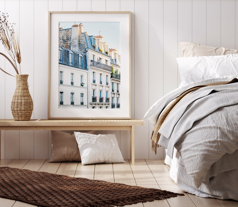 Puede incluir: Una fotograf&iacute;a enmarcada de un edificio parisino con paredes blancas y ventanas azules. El edificio tiene varios balcones y est&aacute; rodeado de otros edificios. La foto est&aacute; colgada en una pared blanca sobre un banco de madera.