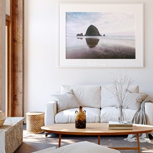 May include: A white sofa with a throw blanket and a vase of white branches sits in front of a large framed photograph of a rocky island in the ocean. A wooden coffee table with a book and a brown ceramic vase sits in front of the sofa.