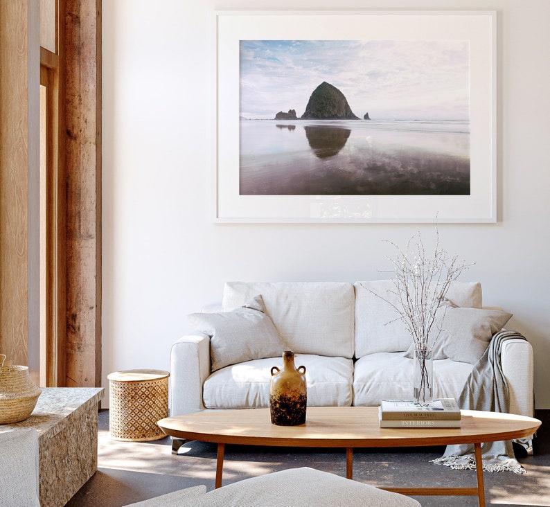May include: A white sofa with a throw blanket and a vase of white branches sits in front of a large framed photograph of a rocky island in the ocean. A wooden coffee table with a book and a brown ceramic vase sits in front of the sofa.