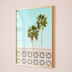 May include: A framed print of two palm trees against a light blue sky. The trees are in front of a white wall with a geometric pattern.