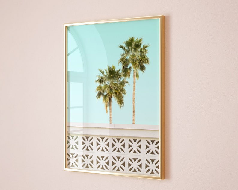May include: A framed print of two palm trees against a light blue sky. The trees are in front of a white wall with a geometric pattern.
