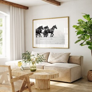 May include: A black and white photograph of a group of black horses running through a field. The photograph is framed in a gold frame and hanging on a white wall above a beige sofa.