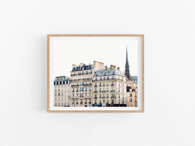 May include: A photograph of a Parisian cityscape with a view of several white buildings with black roofs and windows. The buildings are lined up next to each other and a tall, dark spire is visible in the background.