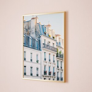 Puede incluir: Una fotograf&iacute;a enmarcada de un edificio parisino con paredes blancas y techos de tejas azul gris&aacute;ceas. El edificio tiene m&uacute;ltiples ventanas y balcones con plantas y flores.