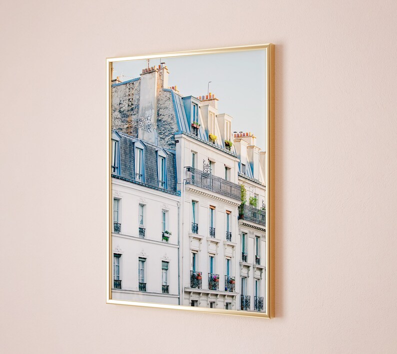 Puede incluir: Una fotograf&iacute;a enmarcada de un edificio parisino con paredes blancas y techos de tejas azul gris&aacute;ceas. El edificio tiene m&uacute;ltiples ventanas y balcones con plantas y flores.