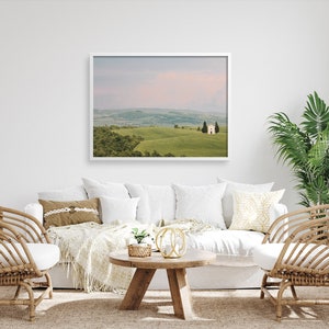 May include: A white framed photograph of a rolling green hill with a small white building on top. The photo is hanging on a white wall above a white couch with brown throw pillows and a white throw blanket. There are two wicker chairs on either side of the couch.