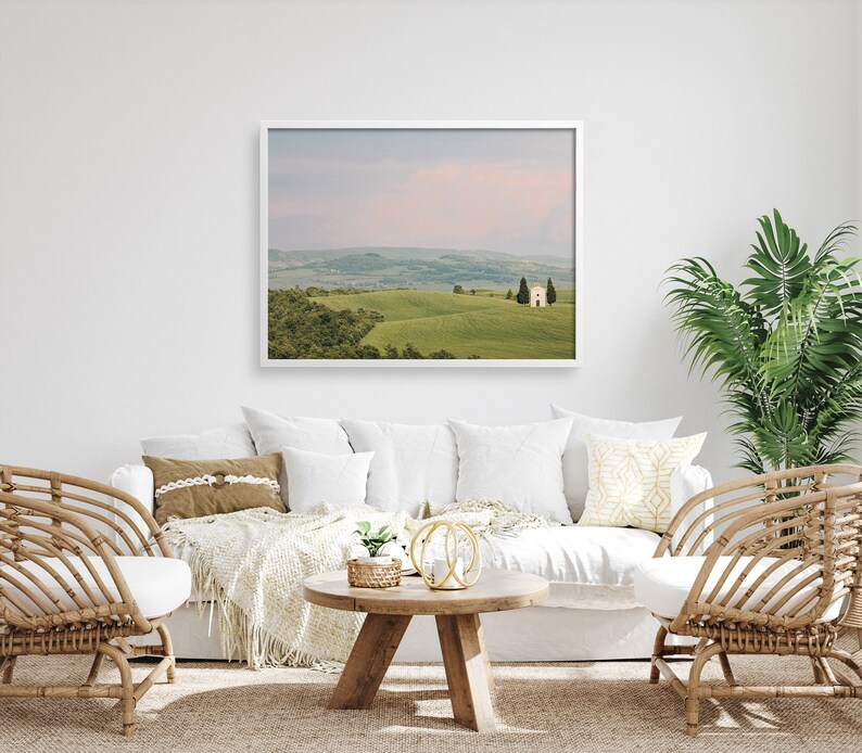 May include: A white framed photograph of a rolling green hill with a small white building on top. The photo is hanging on a white wall above a white couch with brown throw pillows and a white throw blanket. There are two wicker chairs on either side of the couch.