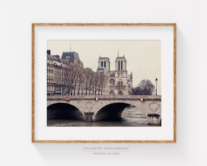 May include: A black and white photograph of the Notre Dame Cathedral in Paris, France. The cathedral is seen from across the Seine River, with a bridge in the foreground. The bridge has stone arches and is lined with trees. The sky is overcast and the photo has a vintage feel.