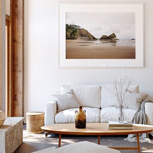 May include: A white couch with two pillows and a throw blanket sits in front of a coffee table with a vase of white branches, a book, and a brown ceramic vase. A framed photograph of a beach scene hangs on the wall behind the couch.