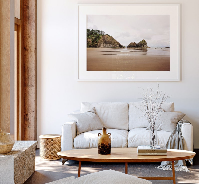 May include: A white couch with two pillows and a throw blanket sits in front of a coffee table with a vase of white branches, a book, and a brown ceramic vase. A framed photograph of a beach scene hangs on the wall behind the couch.