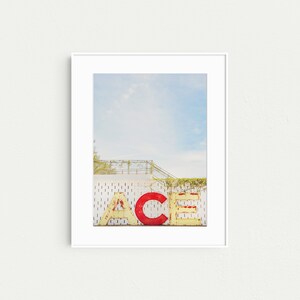 May include: A white framed photograph of a vintage sign that reads "ACE" in large, yellow and red letters. The sign is attached to a white building with a metal railing and green vines.