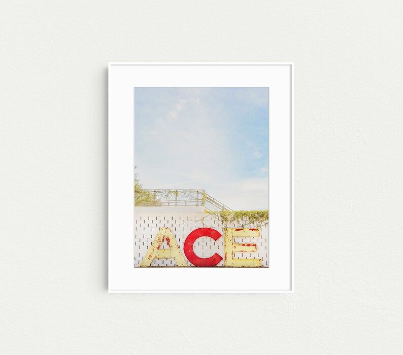 May include: A white framed photograph of a vintage sign that reads "ACE" in large, yellow and red letters. The sign is attached to a white building with a metal railing and green vines.