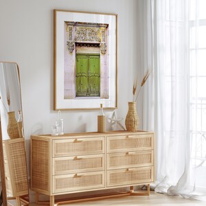 May include: A framed photograph of a green wooden door with ornate carvings above it. The door is set against a pink wall with a white trim. The photo is hanging on a white wall above a wooden dresser with six drawers.