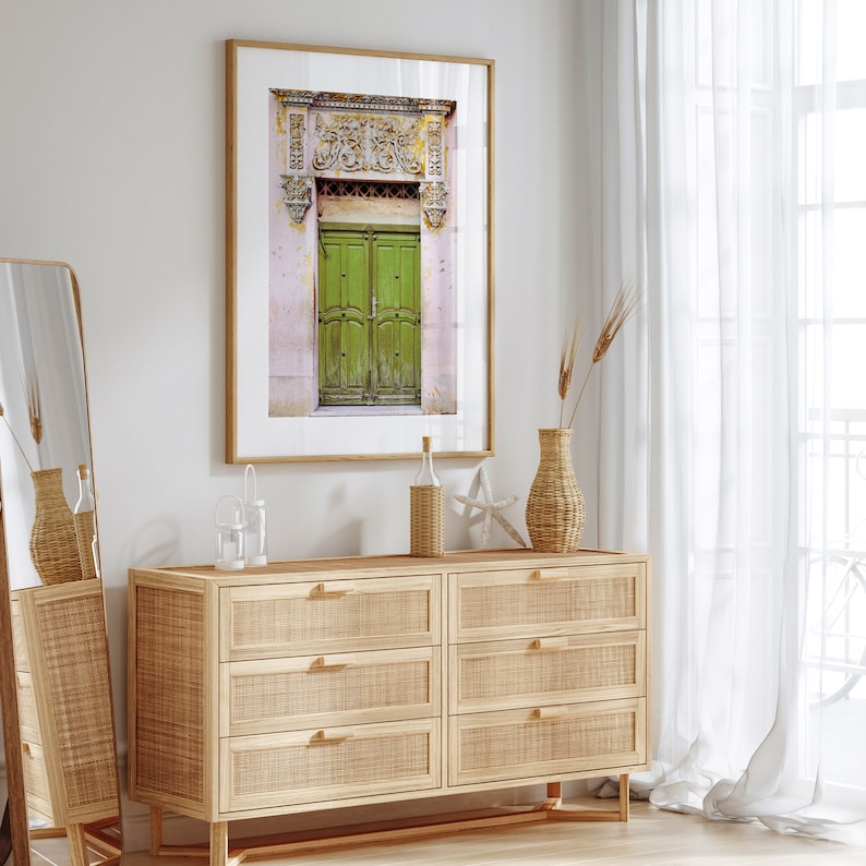 May include: A framed photograph of a green wooden door with ornate carvings above it. The door is set against a pink wall with a white trim. The photo is hanging on a white wall above a wooden dresser with six drawers.