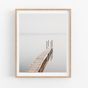 May include: A framed photograph of a wooden dock extending into a still body of water. The dock is made of weathered wood planks and has a few wooden posts supporting it. The water is a light gray color and the sky is a hazy white.