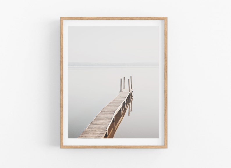 May include: A framed photograph of a wooden dock extending into a still body of water. The dock is made of weathered wood planks and has a few wooden posts supporting it. The water is a light gray color and the sky is a hazy white.