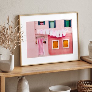 Peut inclure: Une photographie encadr&eacute;e d'un b&acirc;timent rose avec du linge blanc suspendu &agrave; une ligne &agrave; l'ext&eacute;rieur des fen&ecirc;tres. Le b&acirc;timent a deux fen&ecirc;tres avec des volets jaunes et une porte avec un auvent ray&eacute; rose et blanc.