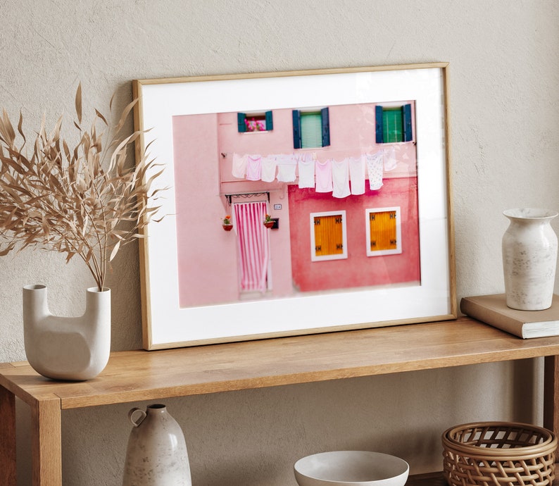 Peut inclure: Une photographie encadr&eacute;e d'un b&acirc;timent rose avec du linge blanc suspendu &agrave; une ligne &agrave; l'ext&eacute;rieur des fen&ecirc;tres. Le b&acirc;timent a deux fen&ecirc;tres avec des volets jaunes et une porte avec un auvent ray&eacute; rose et blanc.