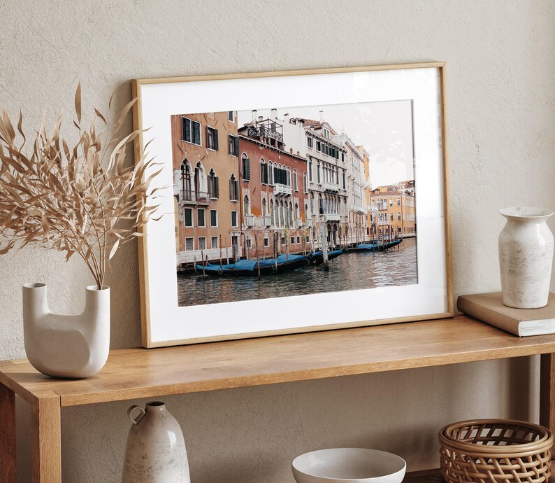 May include: A framed photograph of a canal in Venice, Italy. The photo shows a row of colorful buildings along the canal, with gondolas docked in the water.