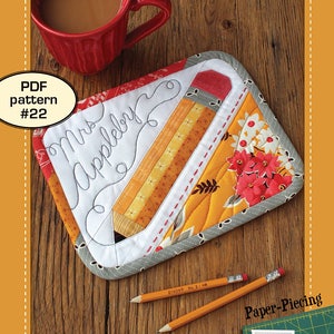 Può includere: Un sottobicchiere a forma di matita trapuntato con il testo "Mrs. Appleby" in corsivo. Il sottobicchiere è realizzato in tessuto patchwork in tonalità di giallo, rosso e grigio. Il sottobicchiere è circondato da due matite gialle e un tappetino da taglio verde con un taglierino rotativo.