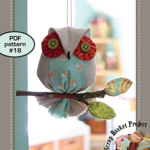 May include: A patchwork owl on a stick, with a green leaf, hanging from a string. The owl is made of fabric and has red and green buttons for eyes. The background is blurred and shows a wooden table and a basket of fabric scraps. The text "a Scrap Basket Project" is written in a circle around the basket. The text "OWL on a STICK" is written below the owl. The text "PDF pattern #18" is in a circle in the top left corner.