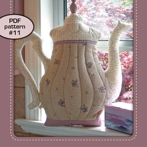 May include: A beige teapot-shaped pillow with a pink trim and floral print. The pillow is sitting on a windowsill with a box of tea behind it. The text "PATCHWORK*POTTERY" and "TEAPOT PILLOW" are visible on the image. The text "PDF pattern #11" is also visible.