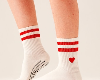 May include: A pair of white athletic socks with red stripes at the top and a red heart graphic. The socks have a ribbed texture and black grip dots on the soles.
