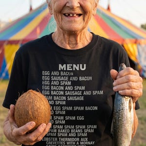 Könnte beinhalten: Schwarzes T-Shirt mit einem Menü von Speisen, das in Weiß gedruckt ist. Das Menü enthält Artikel wie Eier und Speck, Spam und Hummer Thermidor. Eine Kokosnuss und ein Fisch werden vor dem T-Shirt gehalten.